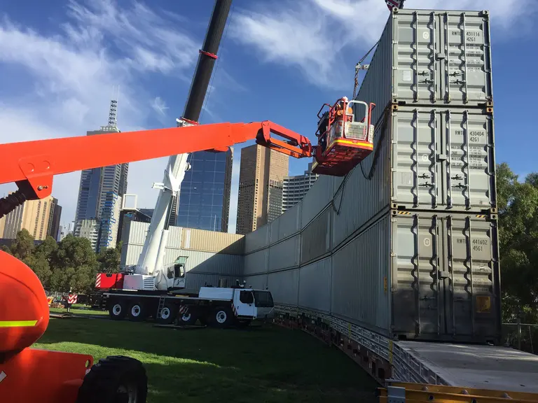 A worker in a cherry picker next to stacked shipping containers, with a large crane and city skyline in the background, indicating temporary worksite facilities.