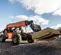 An orange and black JLG SkyTrak telehandler lifts a large stack of plywood on its forks at a construction site, with a residential building visible in the background under a partly cloudy sky.