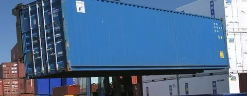 A large yellow forklift lifting a blue shipping container onto a flatbed trailer in a busy container yard.