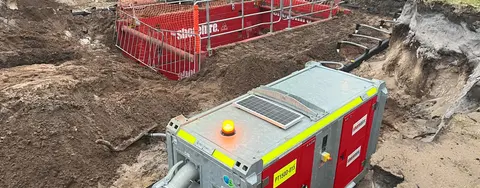 A large red and grey industrial pump with hoses operating in a muddy excavation site with trench shoring.