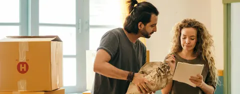 A couple packing or unpacking moving boxes, with one person holding crumpled paper and the other checking a list, suggesting a relocation process.