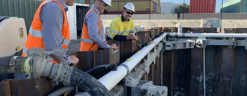 Three construction workers inspecting shoring and piping within a trench at an industrial site with shipping containers.