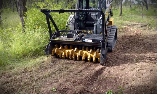 Large machine trimming overgrown grass in an open field near highway barriers to support roadside safety and visibility.
