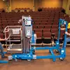 Side view of a blue Genie straddle kit for pusharund lift with arm retracted, idle on a stage in an auditorium.