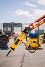 Excavator with a drill attachment at the tip, used for breaking ground at a construction site with safety measures in place.