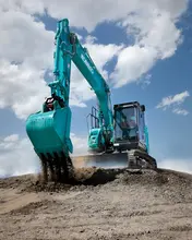 Operator driving an excavator digging soil at a construction site with safety barriers in the background.